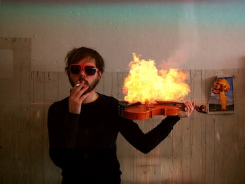 Un hombre barbudo con gafas de sol rojas y camiseta negra fuma un cigarrillo, sosteniendo un violín en llamas, con un cuadro de explosión nuclear detrás