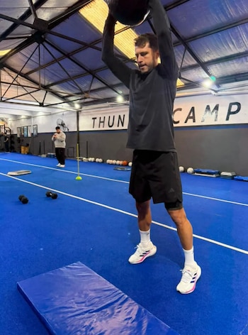 Guido Rodríguez, con camiseta y pantalones cortos negros, levanta una pelota medicinal sobre su cabeza en un gimnasio con suelo azul y cartel "THUN CAMP"