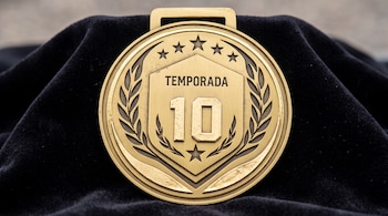 Medalla circular de color dorado con la palabra TEMPORADA y el número 10 en relieve sobre un fondo de tela negra.
