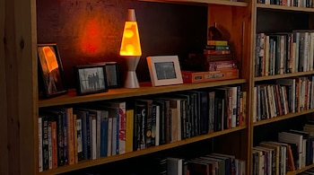 Una estantería de madera clara llena de libros de distintos tamaños, junto a una lámpara de lava naranja encendida, fotos enmarcadas y juegos de mesa.