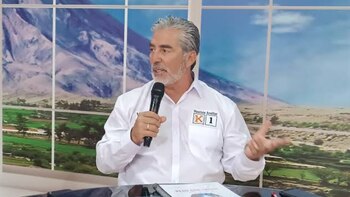Hombre de mediana edad con cabello gris y barba, con camisa blanca y distintivo de campaña, sostiene micrófono. Fondo con imagen de montañas y campos