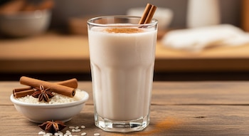 Un vaso de horchata con canela en rama y en polvo, junto a un tazón de arroz blanco, ramas de canela y anís estrellado sobre una mesa de madera.