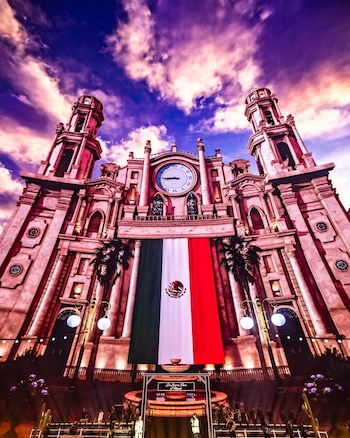 Proyección de una iglesia con la bandera de México y un reloj central sobre un escenario con músicos y farolas
