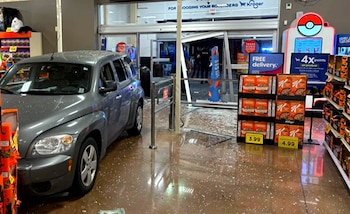 Un automóvil gris dentro de un supermercado rodeado de vidrios rotos junto a la puerta de entrada dañada