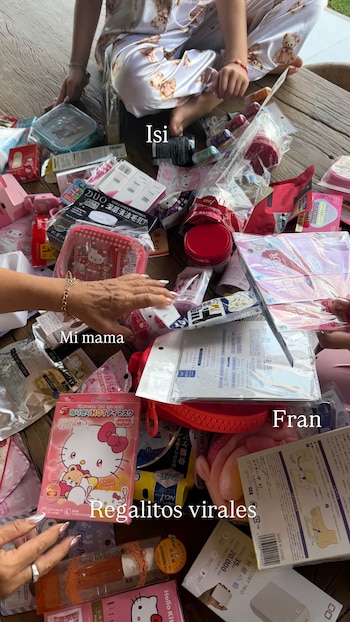 Vista aérea de manos de dos adultos y piernas de una niña rodeadas de numerosos productos de belleza y artículos Hello Kitty sobre un suelo de madera, desempacando regalos