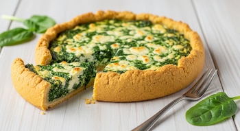 Una tarta redonda de masa dorada con una porción separada, relleno verde de espinaca y ricota, tenedor de metal y hojas frescas de espinaca en una mesa de madera clara.