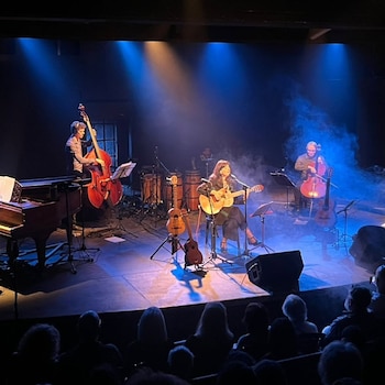 Escenario oscuro con tres músicos, instrumentos como guitarra, contrabajo, violonchelo, piano, batería y micrófono, frente a un público sentado