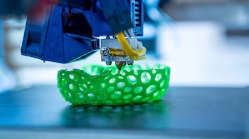 Cabezal de impresora 3D extrayendo filamento verde sobre una estructura esférica perforada del mismo color en una base oscura.
