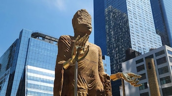 Una gran escultura de madera con manos doradas se alza en primer plano frente a rascacielos de cristal y el cielo azul