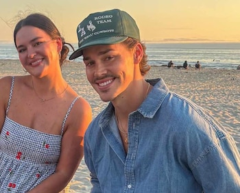 Noah Beck y su hermana sonríen a cámara en una playa durante el atardecer. Él lleva una gorra verde y camisa vaquera; ella, un vestido de cuadros