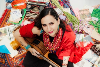 Vista cenital de una mujer con chal rojo y collares que sostiene una guitarra acústica rodeada de mantas, flores y una máquina de escribir