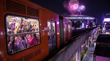 Vagón del Metro de CDMX con fans. Ellos miran fuegos artificiales rosas y violetas en el cielo nocturno sobre el Estadio GNP Seguros.