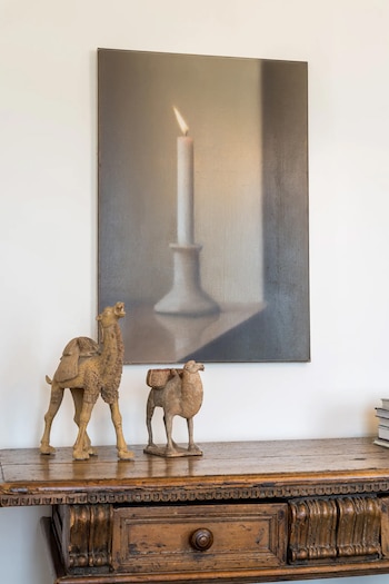 Una pintura vertical de una vela encendida sobre un candelabro se exhibe en una pared blanca, con dos figuras de camellos y una mesa de madera antigua debajo