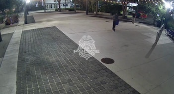 Un hombre con mochila oscura camina por una plaza de adoquines en Miami Beach por la noche, visiblemente un video de vigilancia con el logo de la policía