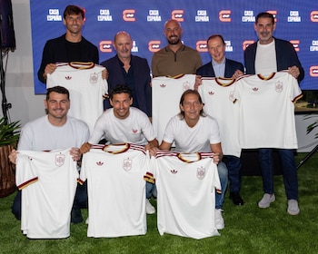 Siete hombres sonrientes sostienen siete camisetas blancas de la selección española de fútbol con detalles rojos y amarillos, ante un panel azul Casa España