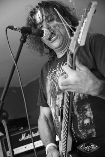 Hombre con pelo largo y rizado tocando una guitarra eléctrica y cantando en un micrófono; amplificador Marshall al fondo. La imagen está en blanco y negro
