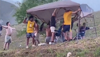Un grupo de jóvenes sujeta una carpa de lona beige en una ladera cubierta de hierba, con un edificio borroso al fondo y el cielo tormentoso