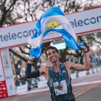 Hombre joven con camiseta de tirantes, bandera de Argentina, medalla en el cuello, brazalete negro y pulseras en la muñeca, sonriendo bajo un arco con logo Telecom