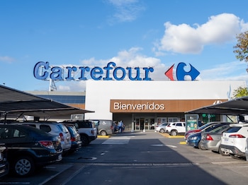 Un edificio blanco de Carrefour con su logo azul y un letrero de "Bienvenidos". Se ve un estacionamiento con muchos coches y un cielo azul con nubes