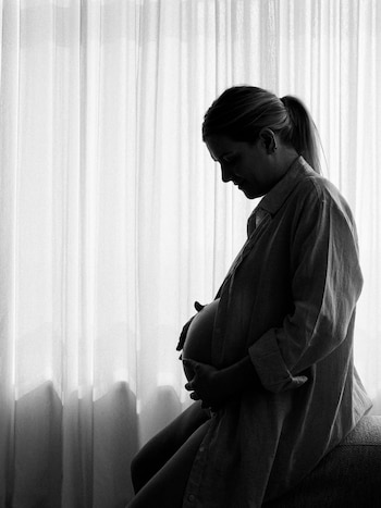 Fotografía en blanco y negro de una mujer sentada de perfil, abrazando su vientre abultado. La mujer tiene el cabello recogido y está frente a una cortina blanca