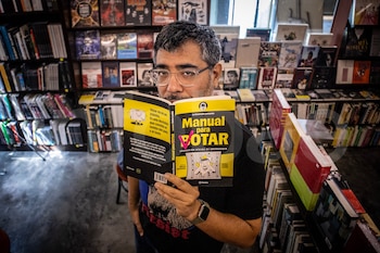 Hombre con gafas y barba sostiene un libro amarillo con el título 'Manual para Votar' frente a su rostro en una librería