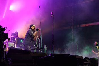 Un cantante barbudo con gorra y guantes canta en un micrófono en un escenario iluminado con luces púrpuras y rosas y humo, con un bajista al fondo