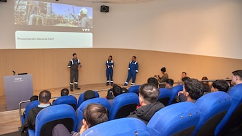 Tres profesionales con uniforme industrial frente a una pantalla de proyección que muestra el logo YPF, texto CILP y una fábrica. Hay una audiencia sentada en sillas azules