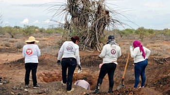 Vista trasera de cinco personas, cuatro mujeres y un hombre, excavando en un terreno desértico de tierra rojiza con arbustos secos y un cielo nublado.