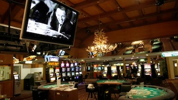 Interior de un casino con mesas de juego, máquinas tragamonedas y un televisor grande mostrando una imagen en blanco y negro de un hombre sonriendo