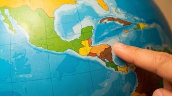 Primer plano de un dedo apuntando a la región de Centroamérica y el Caribe en un globo terráqueo, con tierras verdes, amarillas y mar azul.