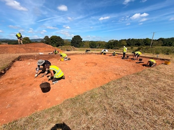 Personas con chalecos reflectantes y sombreros excavan en un terreno rojizo. Hay cubos, palas, herramientas, hierba, árboles y cielo con nubes