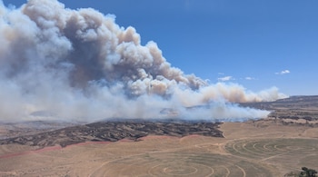 Vasto paisaje desértico con una columna de humo de incendio forestal, terreno quemado negro, una línea roja, campos circulares verdes y cielo azul