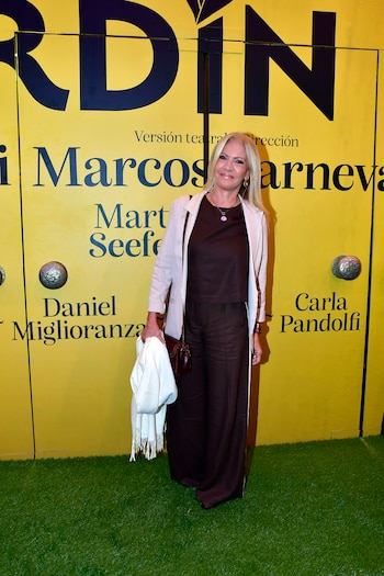 Una mujer rubia sonriente con saco beige y pantalón marrón posa frente a un telón amarillo con el título de una obra y nombres de actores, sobre césped artificial