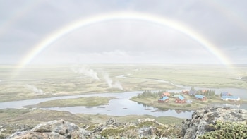 Arcoíris sobre paisaje con rocas, vegetación, río sinuoso, un pueblo con casas de colores y columnas de vapor