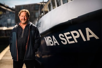 Hombre de mediana edad con barba y chaqueta azul sonríe de pie junto al costado de un barco azul y blanco con el nombre 'MBA SEPIA' en un muelle soleado