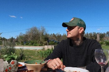 Un hombre con gorra verde, suéter negro y tatuajes en el cuello sentado a una mesa al aire libre frente a un plato y una copa