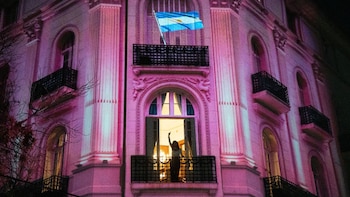 Edificio de estilo clásico con fachada rosa iluminada, bandera argentina proyectada en un balcón superior y una persona saludando desde un balcón inferior