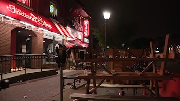Vista nocturna de una calle con un edificio de ladrillo rojo y un toldo rojo. Hay mesas de pícnic volcadas y sillas apiladas en la acera