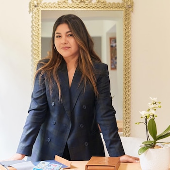Una mujer con cabello largo y saco de cuadros se apoya en una mesa con muestras de tela, un libro y una orquídea. Detrás hay un espejo con marco dorado