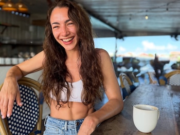 Una mujer de cabello largo y castaño sonríe sentada a una mesa de madera que tiene una taza blanca en un establecimiento abierto