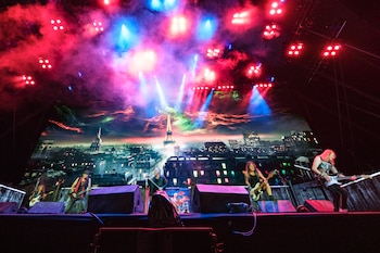 Músicos interpretan canciones con guitarras sobre un escenario iluminado con luces de colores y un fondo de la ciudad de París
