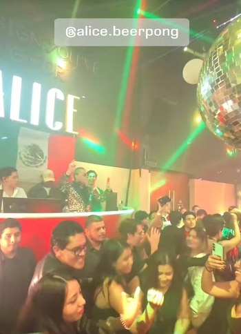 Interior de una discoteca llena de gente bailando bajo luces láser rojas y verdes. Un DJ con una chaqueta brillante está en el escenario y una bola de discoteca cuelga del techo