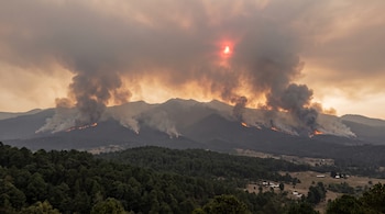 Cordillera montañosa con llamas y grandes columnas de humo que ascienden hacia un cielo cubierto por una capa de humo y un sol rojizo.