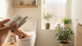 Primer plano de una persona leyendo una revista sentada en el inodoro de un baño moderno con luz natural, plantas, toallas enrolladas y un reloj de arena.
