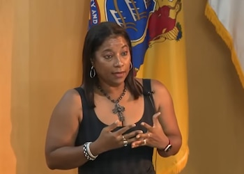 Una mujer con vestido negro, collar con cruz y pendientes gesticula con las manos delante de una bandera