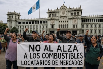 Varias personas con los puños en alto sostienen una pancarta blanca frente a un edificio histórico con la bandera de Guatemala.