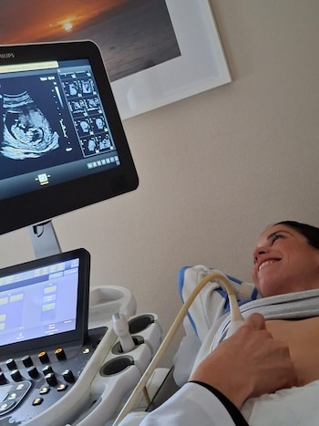Mujer recostada con un monitor de ecografía mostrando la imagen de un feto, una sonda de ecografía en su abdomen y el brazo de un profesional