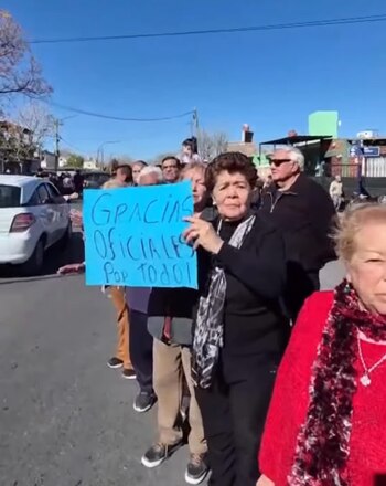 Grupo de personas en una calle, una mujer con un cartel azul que dice 'GRACIAS OFICIALES POR TODO!'. Hay un auto blanco y edificios al fondo bajo cielo azul