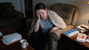 Mujer con expresión de cansancio extremo sentada en un sillón con una manta, su mano en la frente. Sobre una mesa hay un pastillero de Levothyroxine y un folleto de tiroides.