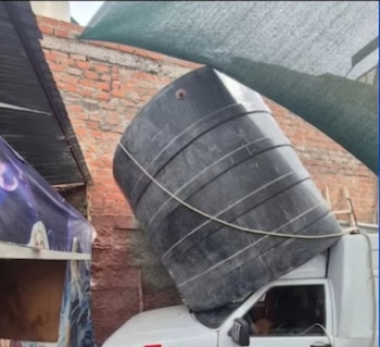Un gran tanque de agua negro está inclinado, apoyado sobre la cabina de una camioneta blanca frente a una pared de ladrillos, con una lona verde arriba